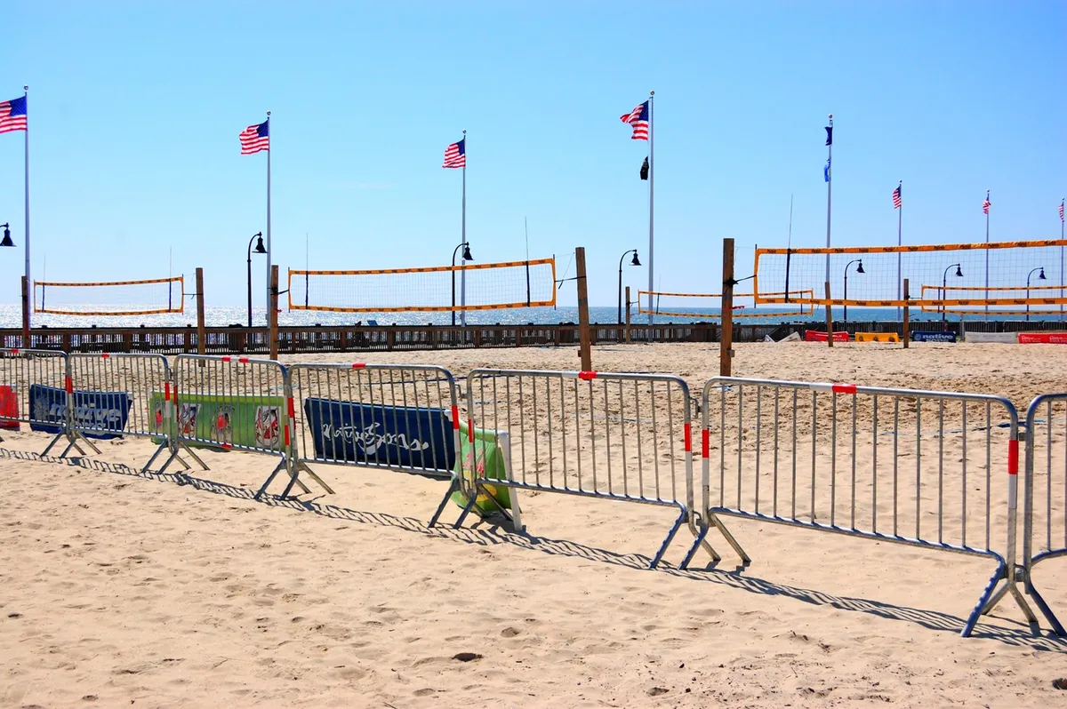 An energetic beach scene with volleyball nets set up in the sand, American flags fluttering, and groups of friends playing or lounging nearby. The open stretch of sand and visible ocean create an inviting backdrop for social activities and relaxation.