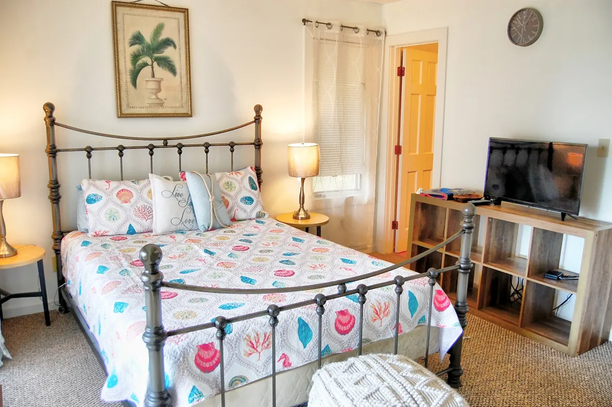 This image features a comfortable bed adorned with a colorful quilt, accompanied by a nightstand with a lamp and a television unit. The space is filled with sunlight, highlighting a warm and cozy atmosphere ideal for rest and relaxation.