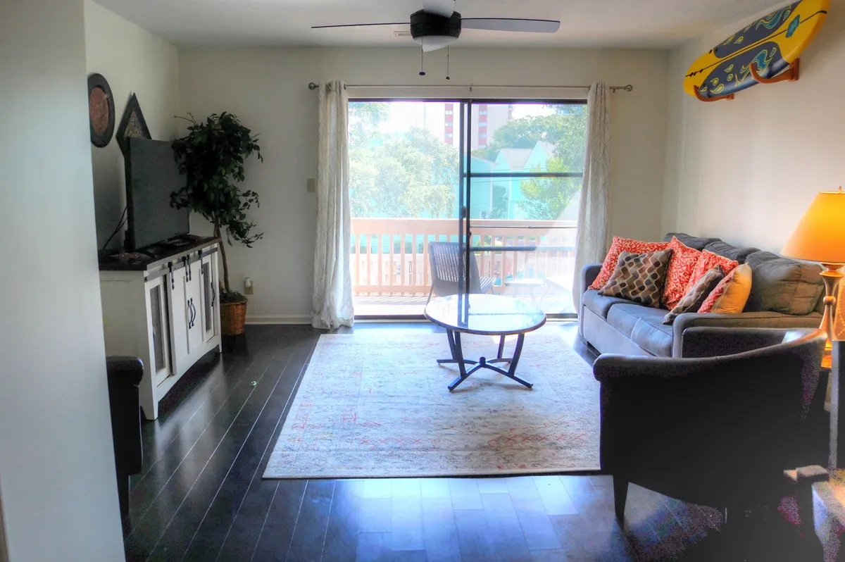 A cozy, sun-filled living area with a sofa adorned with colorful cushions, a coffee table, a television unit, and a large window offering an outside view. The space is styled for both comfort and versatility, perfect for entertaining, unwinding, or enjoying time with family and friends.