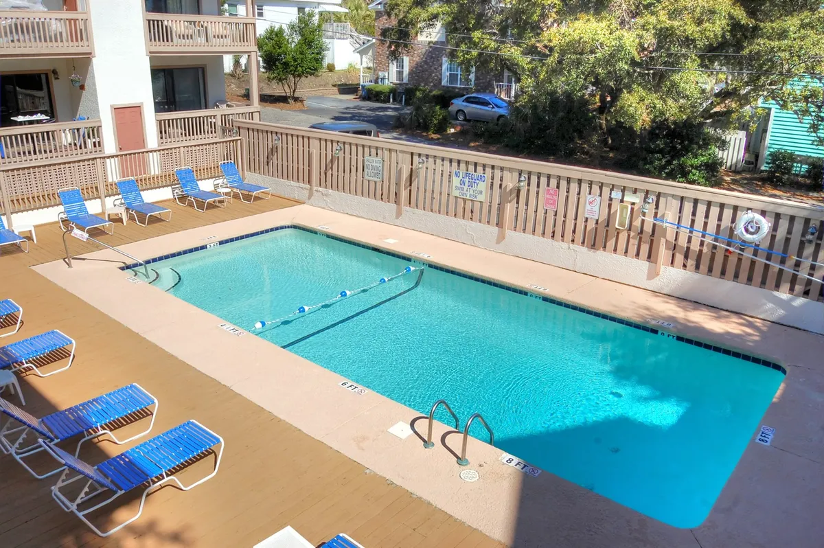 A sparkling outdoor pool is the focal point, surrounded by lounge chairs and shaded by lush trees. The pool area has a spacious deck for sunbathing, socializing, or relaxing in the water, and exudes a resort-style leisure atmosphere.