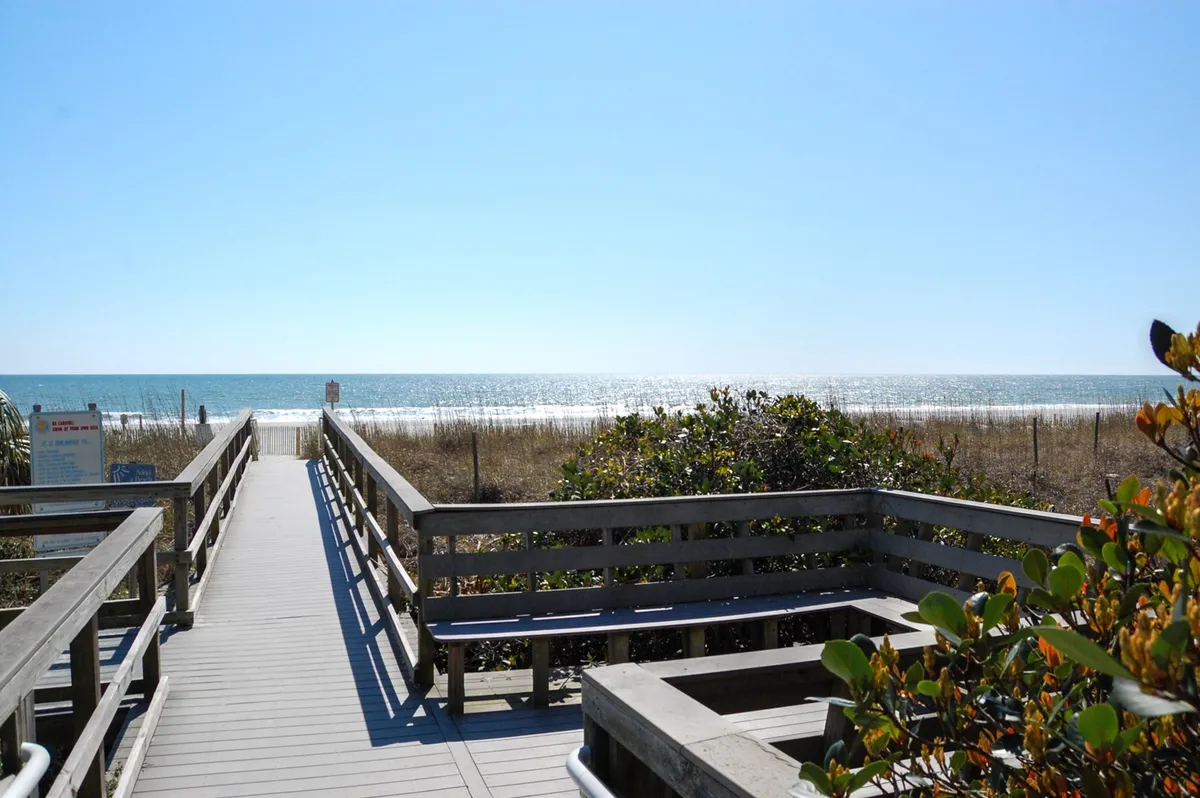 An inviting wooden walkway leads out through sand dunes and vegetation toward the open ocean and distant horizon. The scene is peaceful and emphasizes proximity to the beach, evoking relaxing strolls and easy access to the coast.