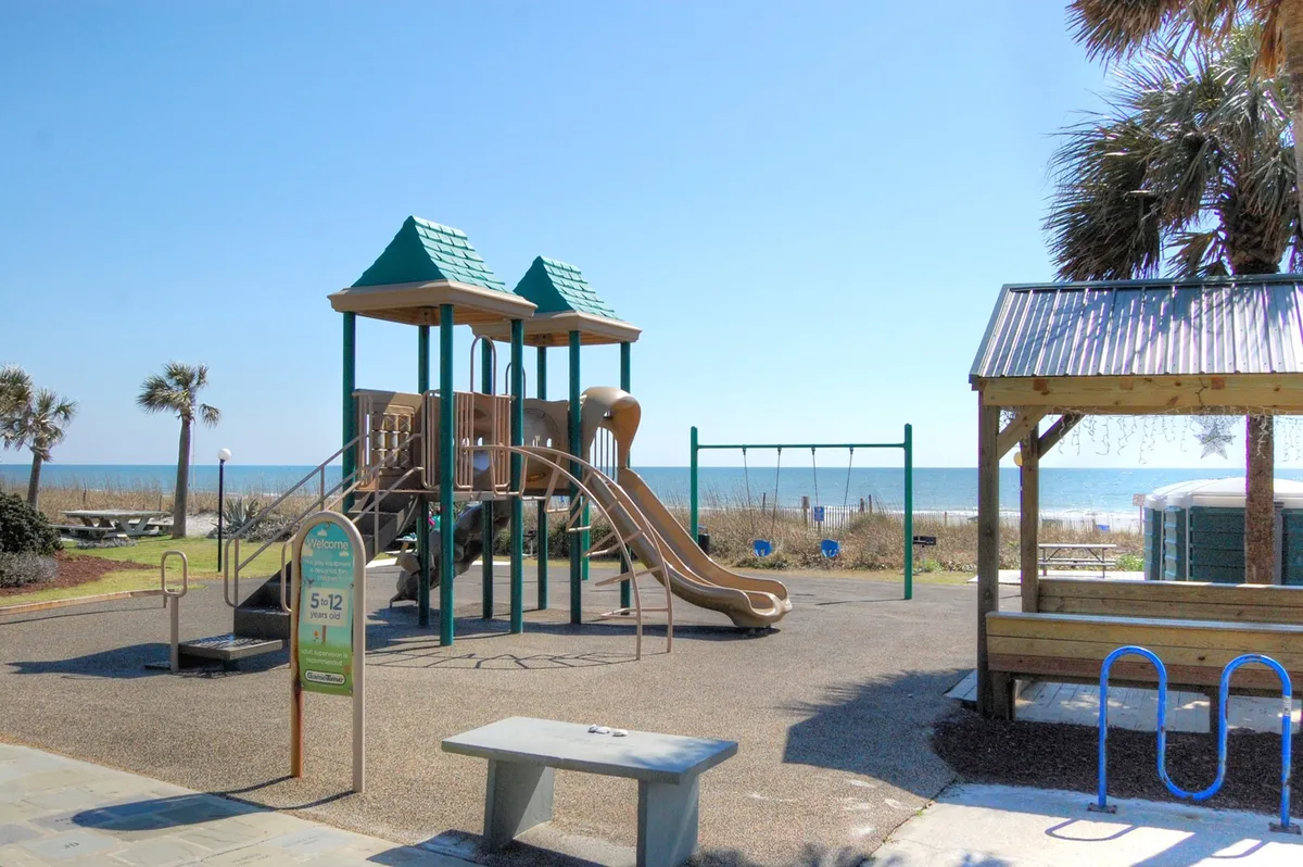 A colorful playground with swings and climbing structures sits just steps from the ocean, framed by palm trees. Children laugh and play under the sunny sky, while families relax nearby, enjoying picnics and the sound of the waves. The backdrop of the beach and water reinforces the family-friendly, carefree atmosphere.