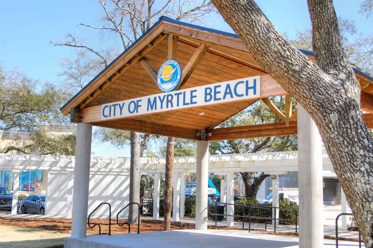 This exterior shot features the recognizable 'City of Myrtle Beach' sign beneath a wooden pavilion structure, surrounded by lush trees. The scene suggests a welcoming environment, indicative of local community engagement and easy access to city amenities.