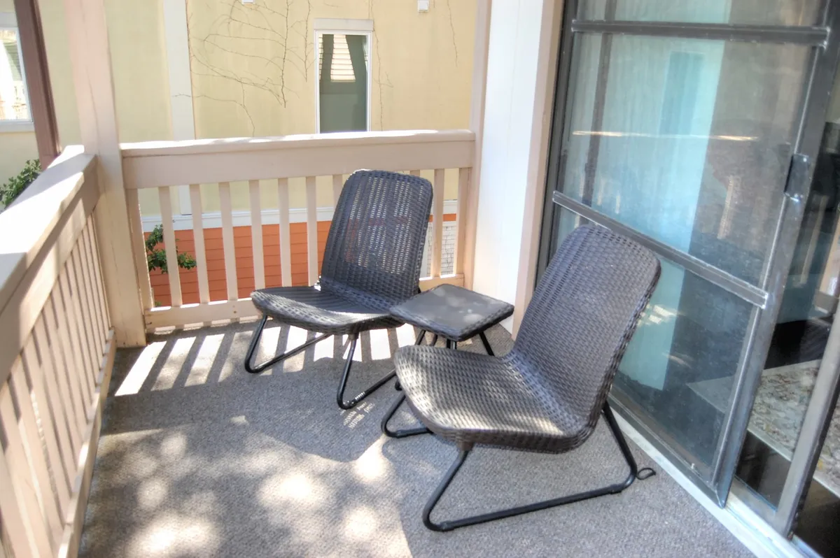 A private outdoor balcony setup featuring two black chairs and a small table, suggesting a cozy spot for unwinding. The setting overlooks lush landscaping, inviting guests to enjoy drinks or share stories in the open air.
