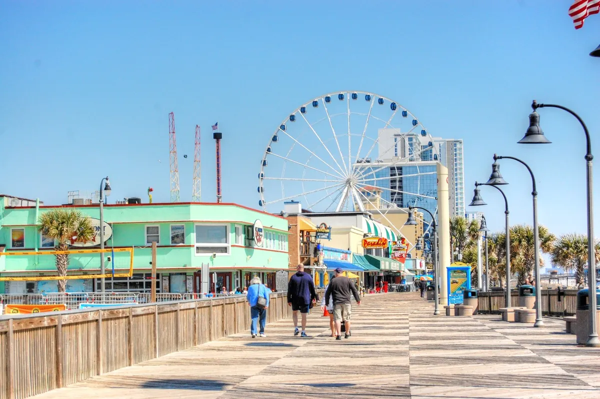 A lively boardwalk scene featuring a brightly lit ferris wheel, colorful storefronts, and palm trees, bustling with activity. Pedestrians stroll along the boardwalk, window shopping and enjoying the festive atmosphere, while dining options and entertainment venues are visible in the background.