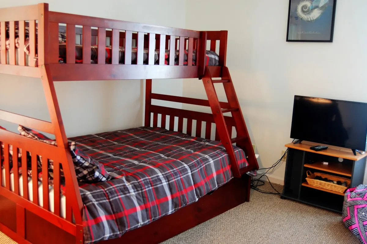 A relaxed bedroom setting with a pyramid bunk bed (double base with a twin top), a television, and plaid bedding. The arrangement offers versatile sleeping options, making it welcoming for kids, friends, or families. The mood is relaxed and functional, supporting memorable sleepovers or movie nights.